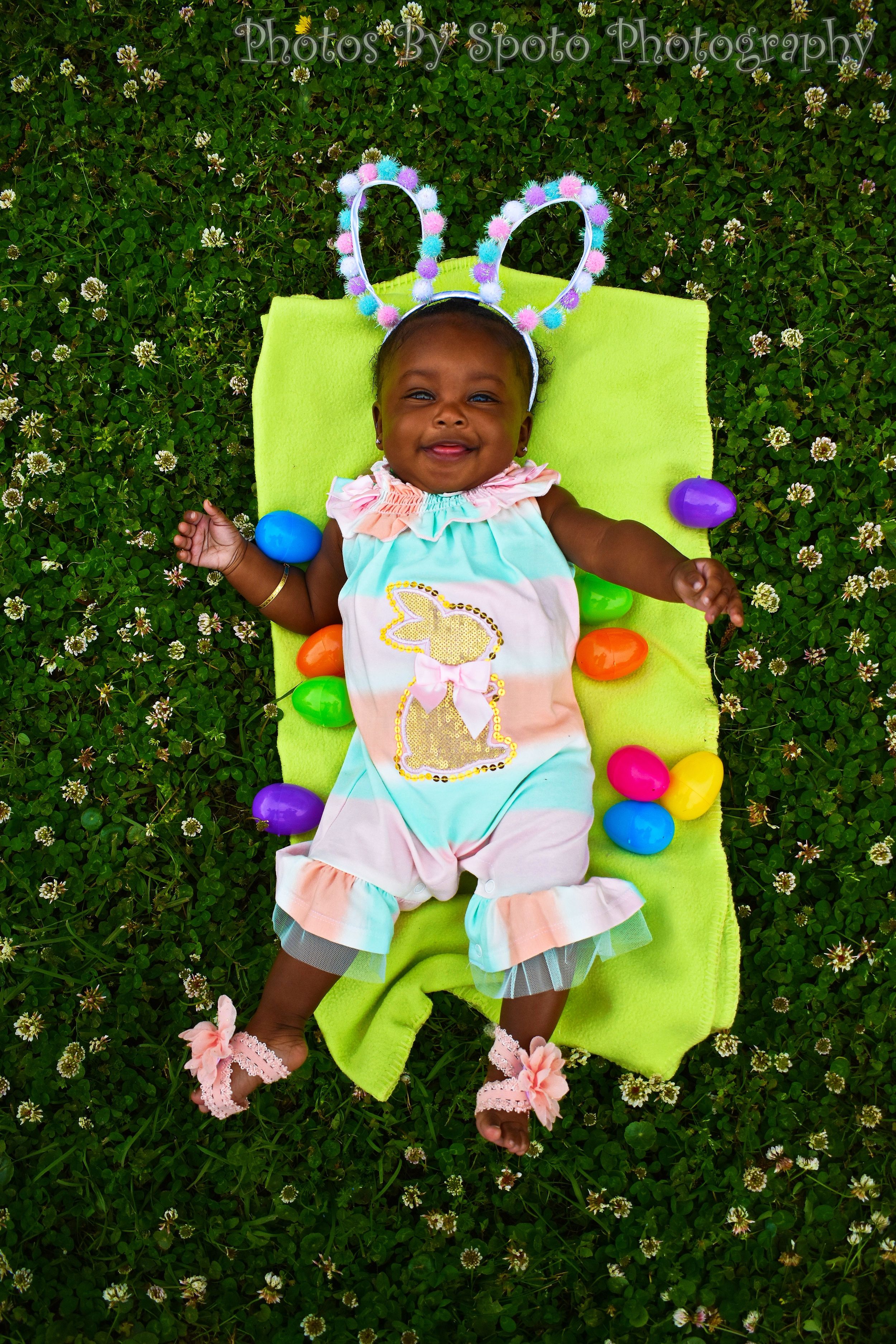 Baby lying on bright green blanket surrounded by colorful plastic Easter eggs and spring flowers in grass.