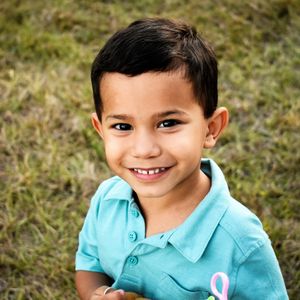 Child wearing light blue polo shirt smiles warmly while sitting outdoors on grass.