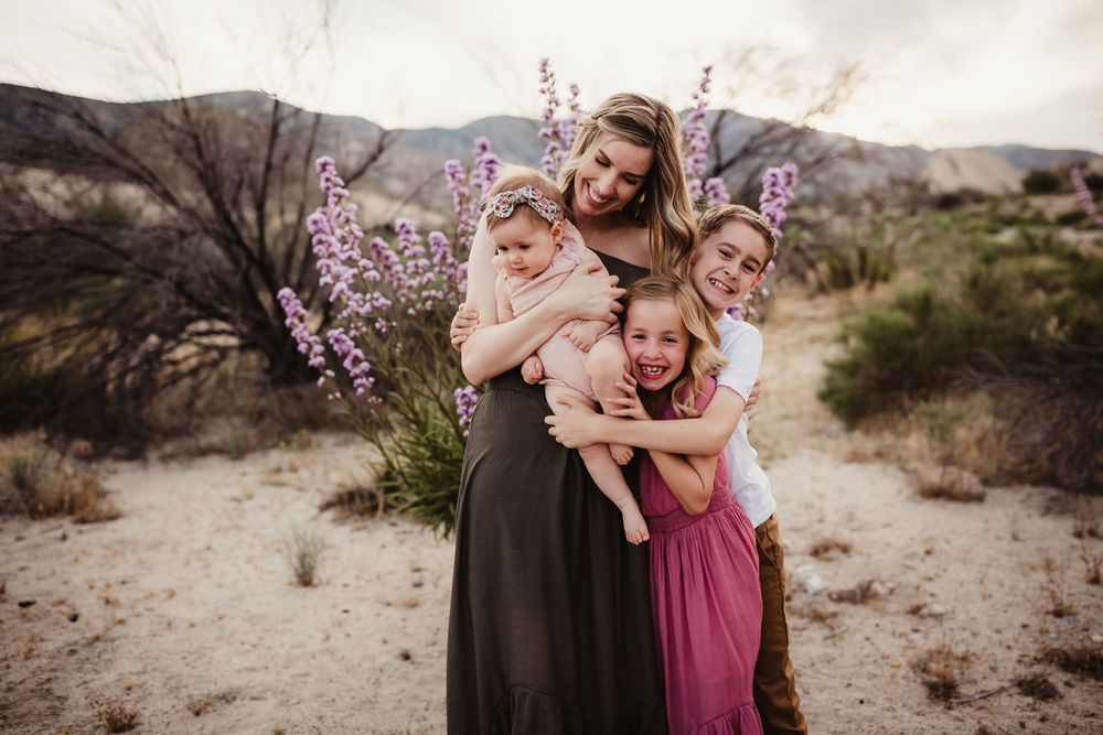 Family embracing in a desert landscape with mountains and purple wildflowers at sunset.