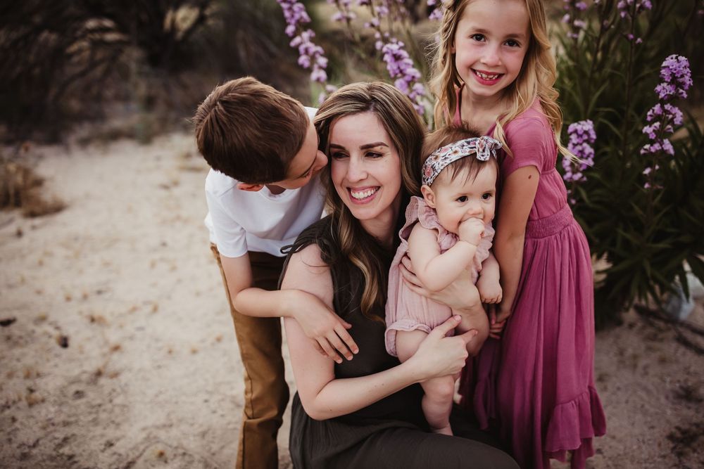 Family in mauve and neutral tones embraces together during outdoor portrait session with purple flowering plants.