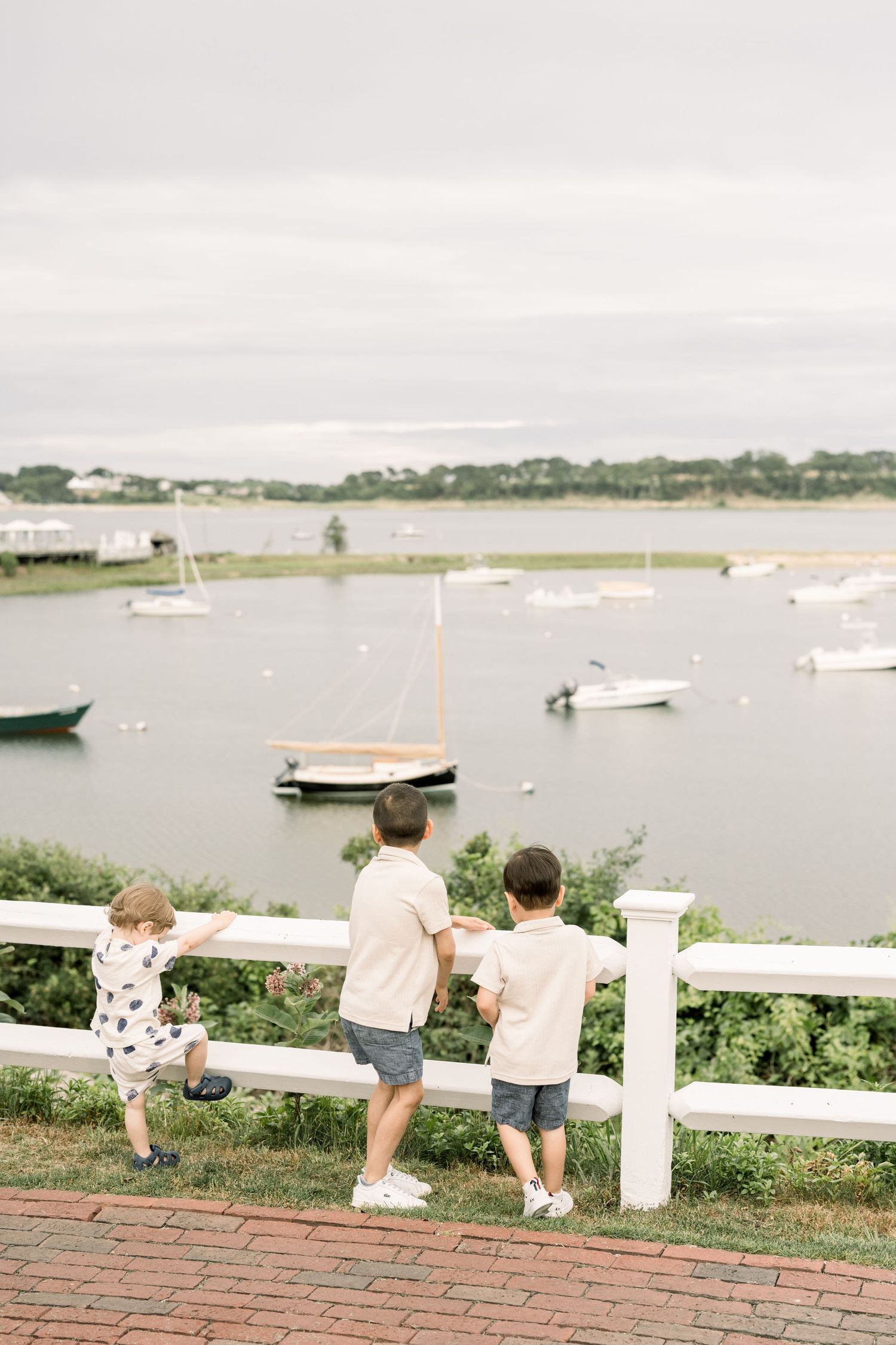 Children lean against a white fence overlooking a harbor with boats on a cloudy summer day.