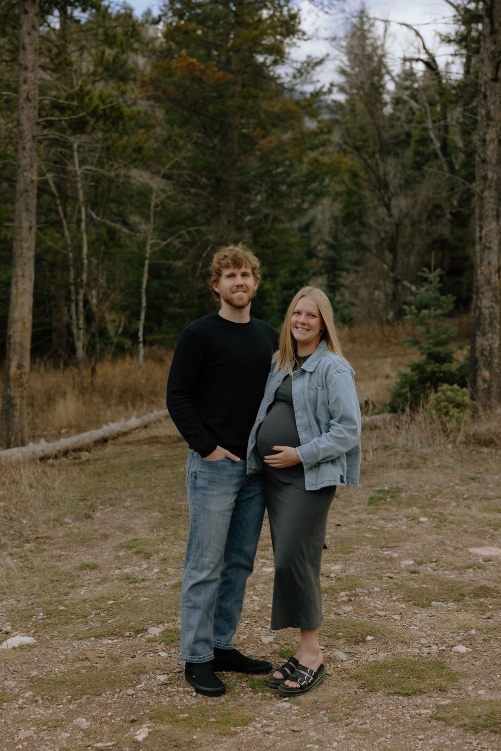 A couple stands together in a wooded outdoor setting wearing casual denim and dark clothing.