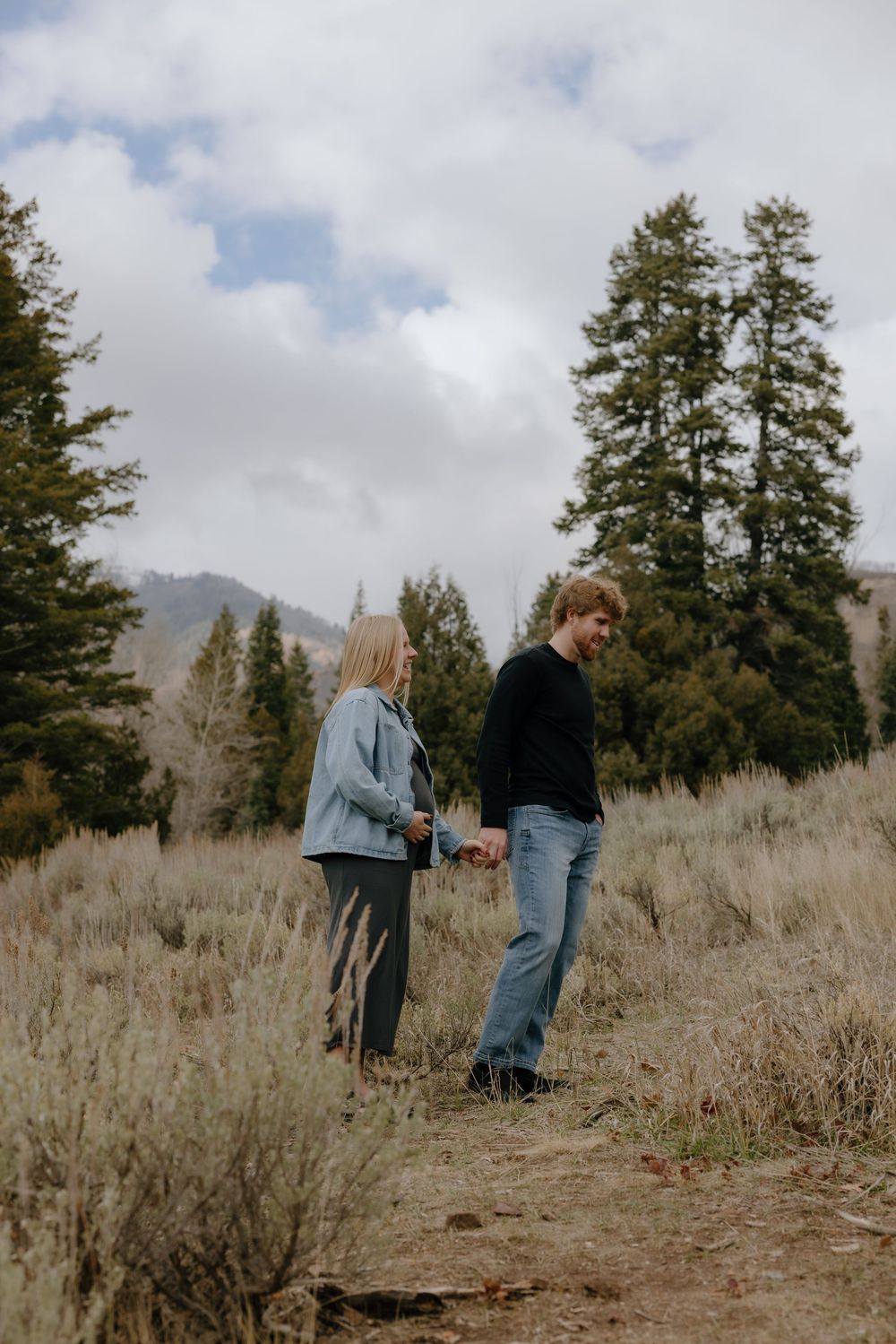 A couple stands together in a field of tall grass with evergreen trees and cloudy skies in the background.