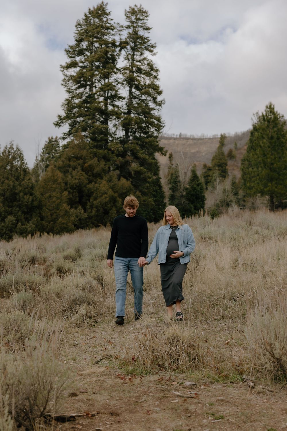 Two people walk together through a grassy hillside with evergreen trees and cloudy skies in the background.