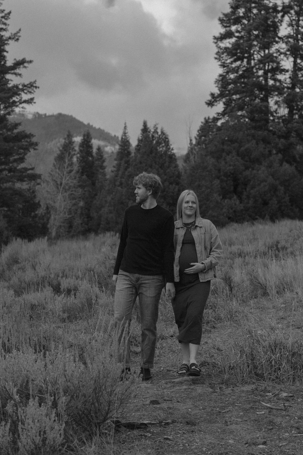 A couple walks through a misty mountain meadow surrounded by tall pine trees in black and white.