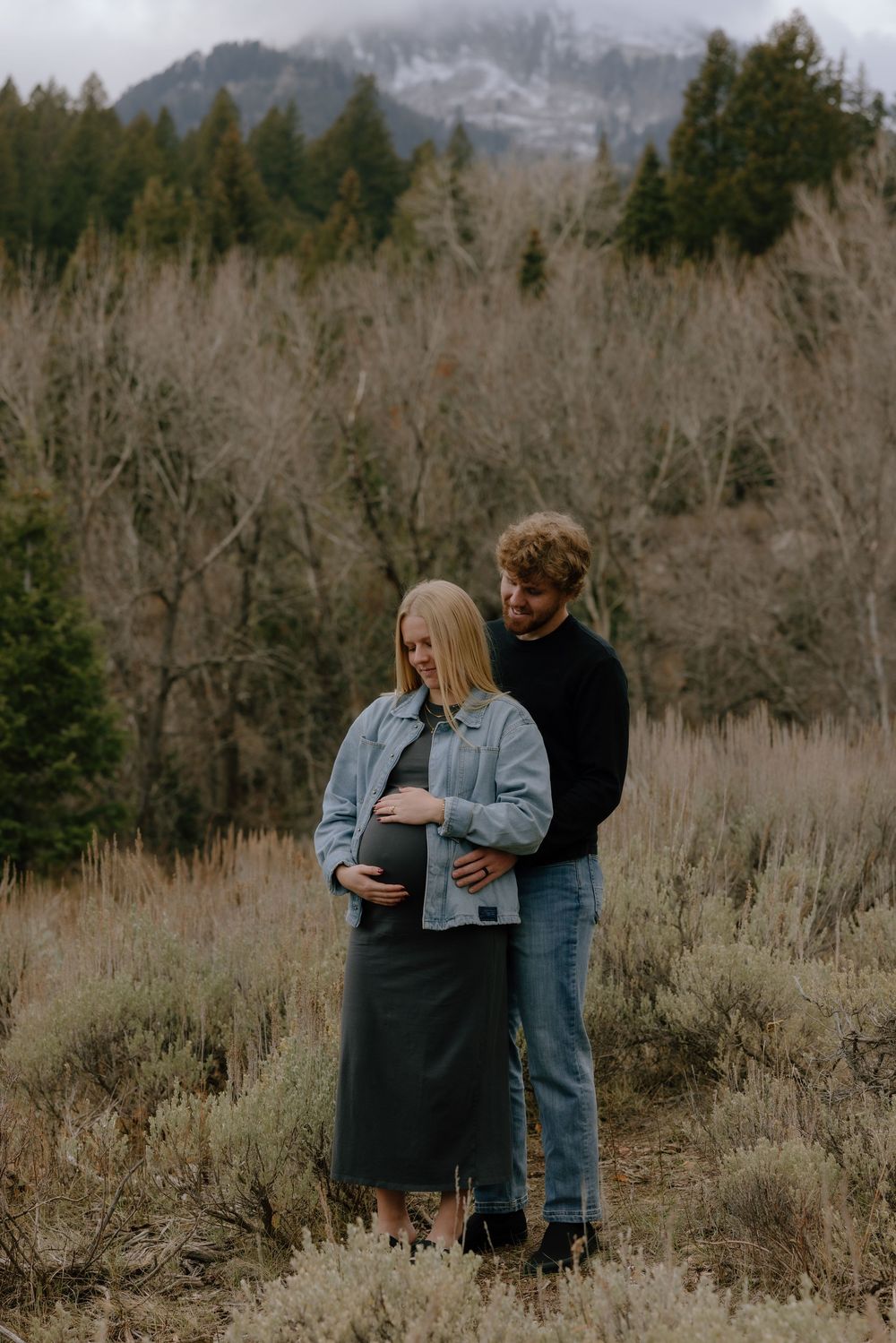 A couple embraces in a scenic mountain landscape with bare trees and natural terrain in the background.