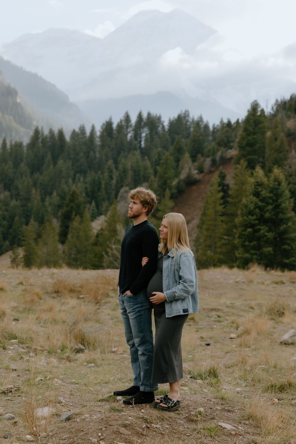 Two people stand together in a mountain meadow surrounded by evergreen forest and misty peaks in the background.