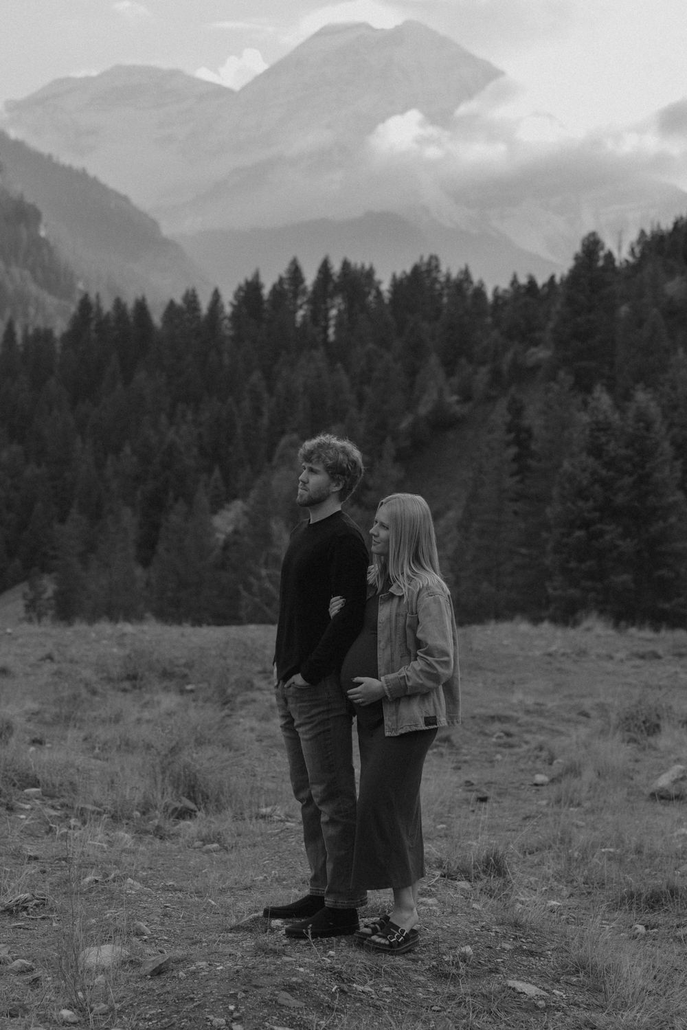 A couple stands in a mountain meadow with evergreen forests and peaks in the background.