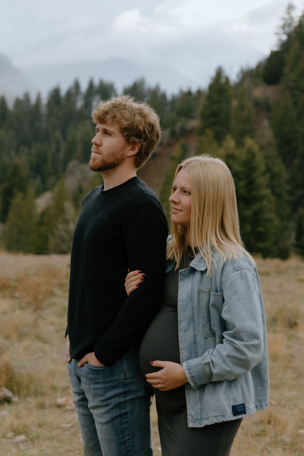 A couple embraces in a field with evergreen trees in the background on an overcast day.