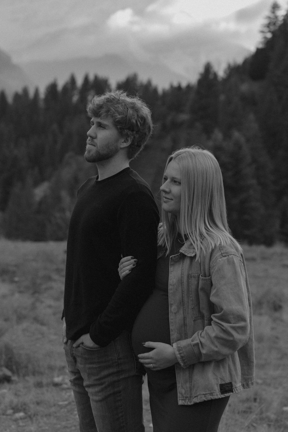 A couple shares intimate moments in a field with mountains and forest in the background.