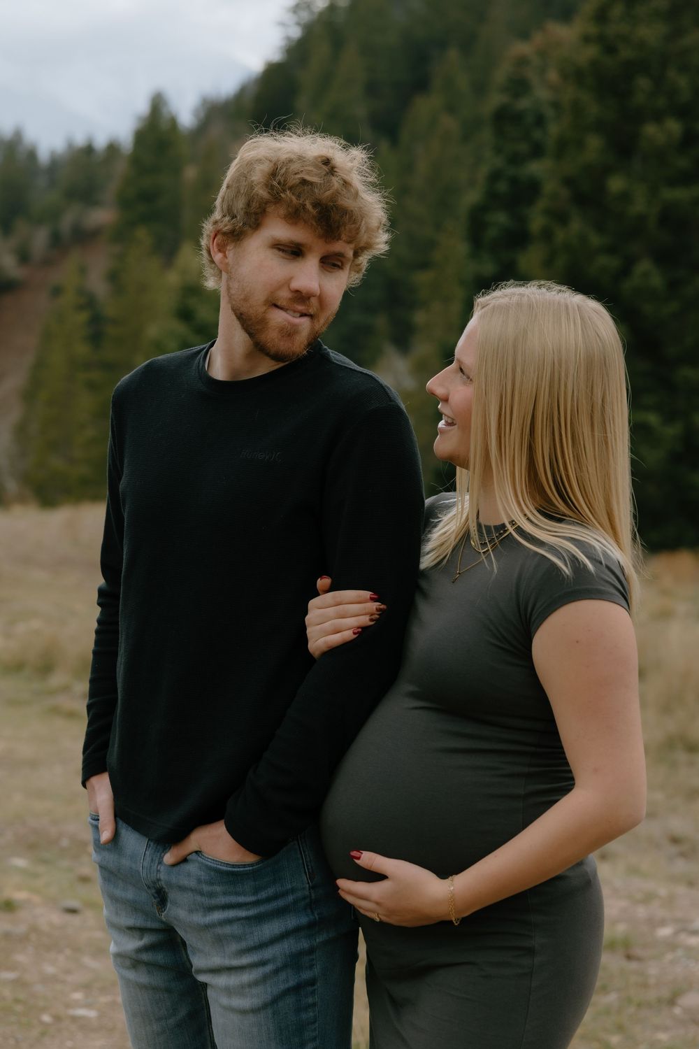 Couple poses outdoors in nature during maternity photoshoot with forest backdrop and moody fall lighting.
