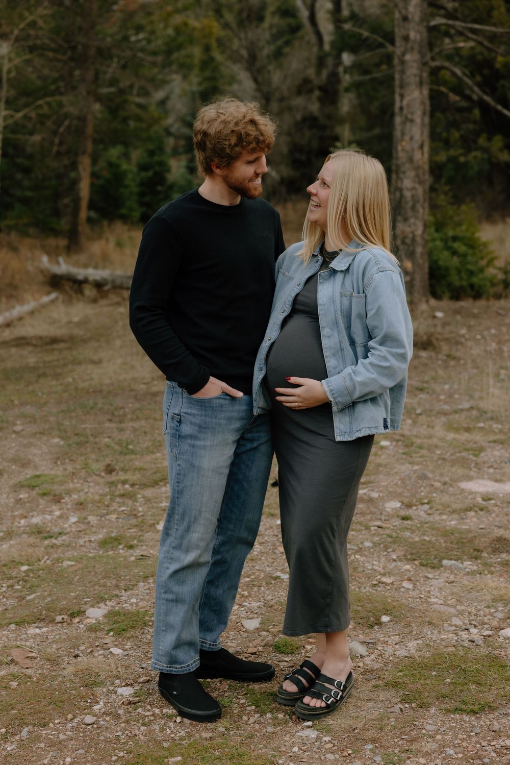A couple in casual denim outfits stands together against a wooded backdrop on rocky ground.