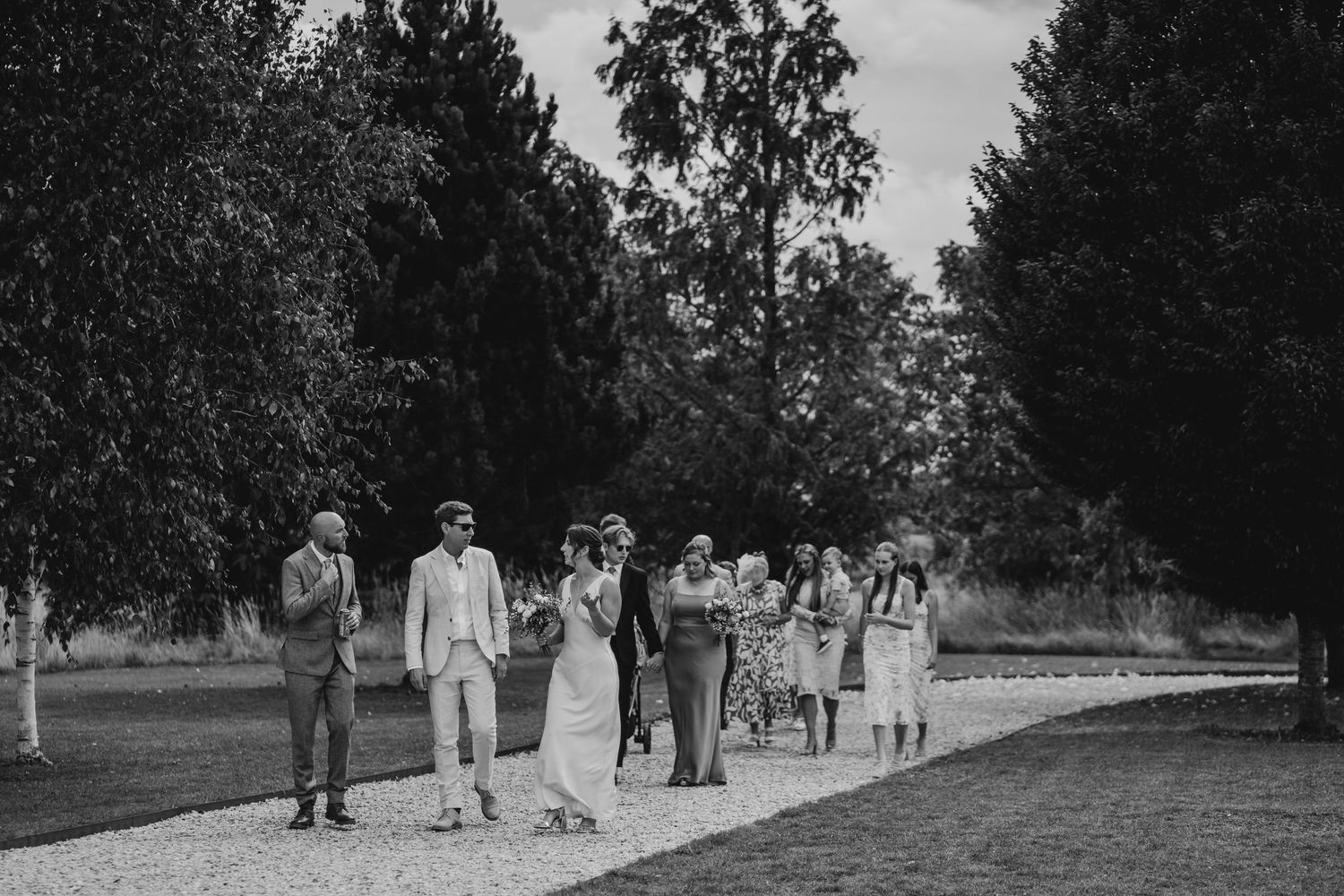 A black and white panoramic photograph shows a group of people walking along a tree-lined path in formal attire.