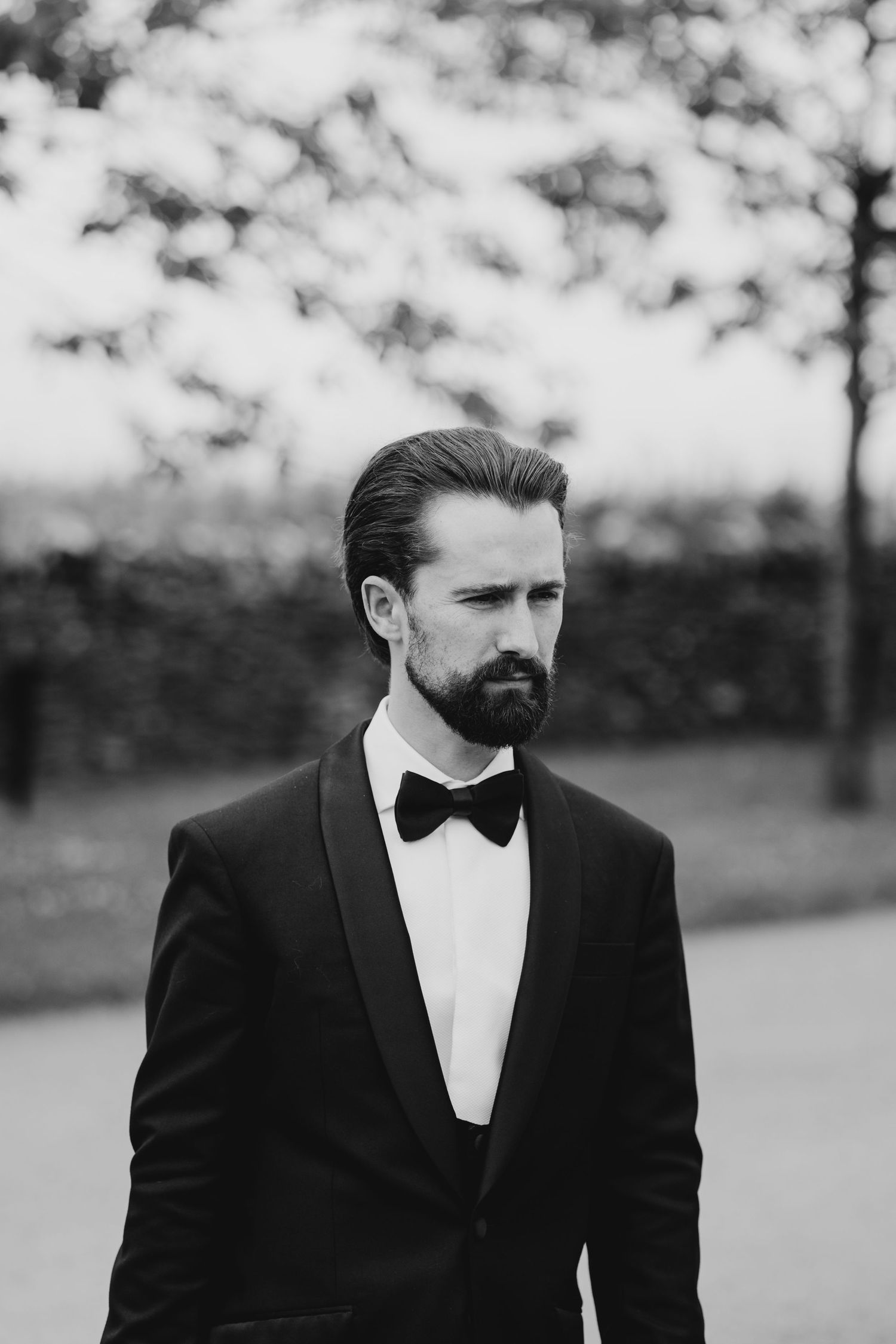 Black and white portrait of a person in a tuxedo and bow tie standing outdoors with a blurred background.