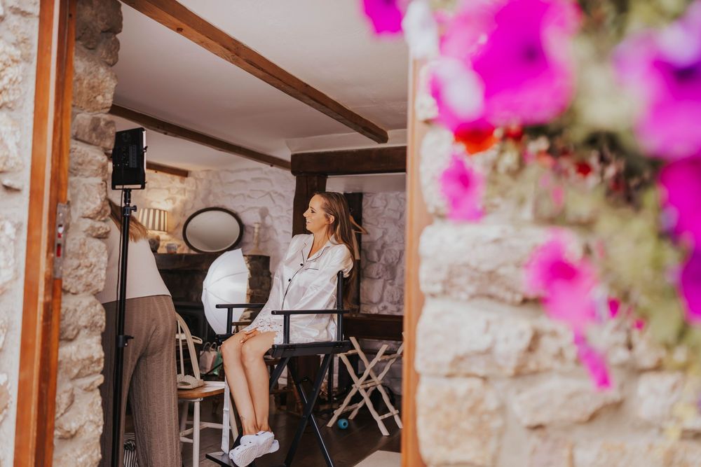 A person in white sits at a makeup station with bright pink flowers in the foreground against a stone wall.