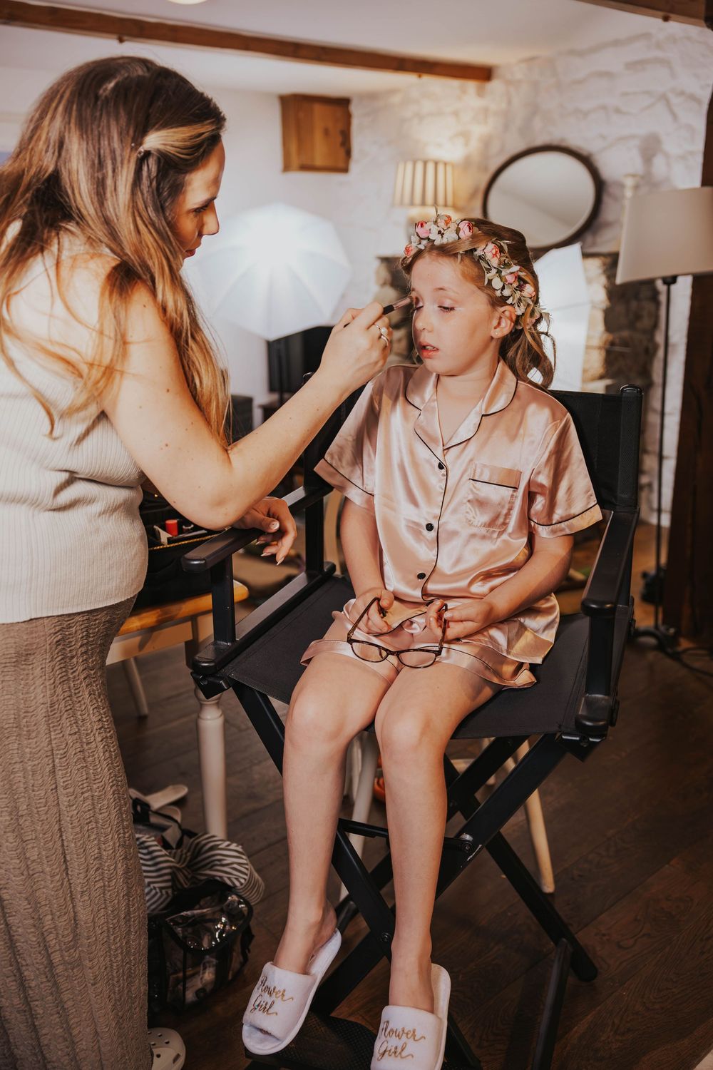 A makeup artist applies cosmetics to a young person wearing silk pajamas and curlers while seated in a director's chair.