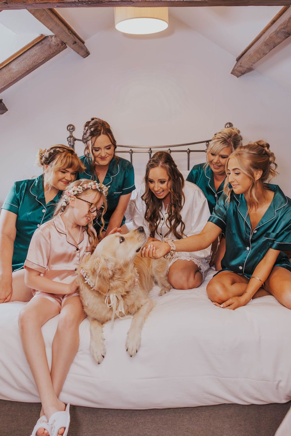 Group in matching teal robes relaxing on white bedding with golden retriever dog during pre-wedding preparations.
