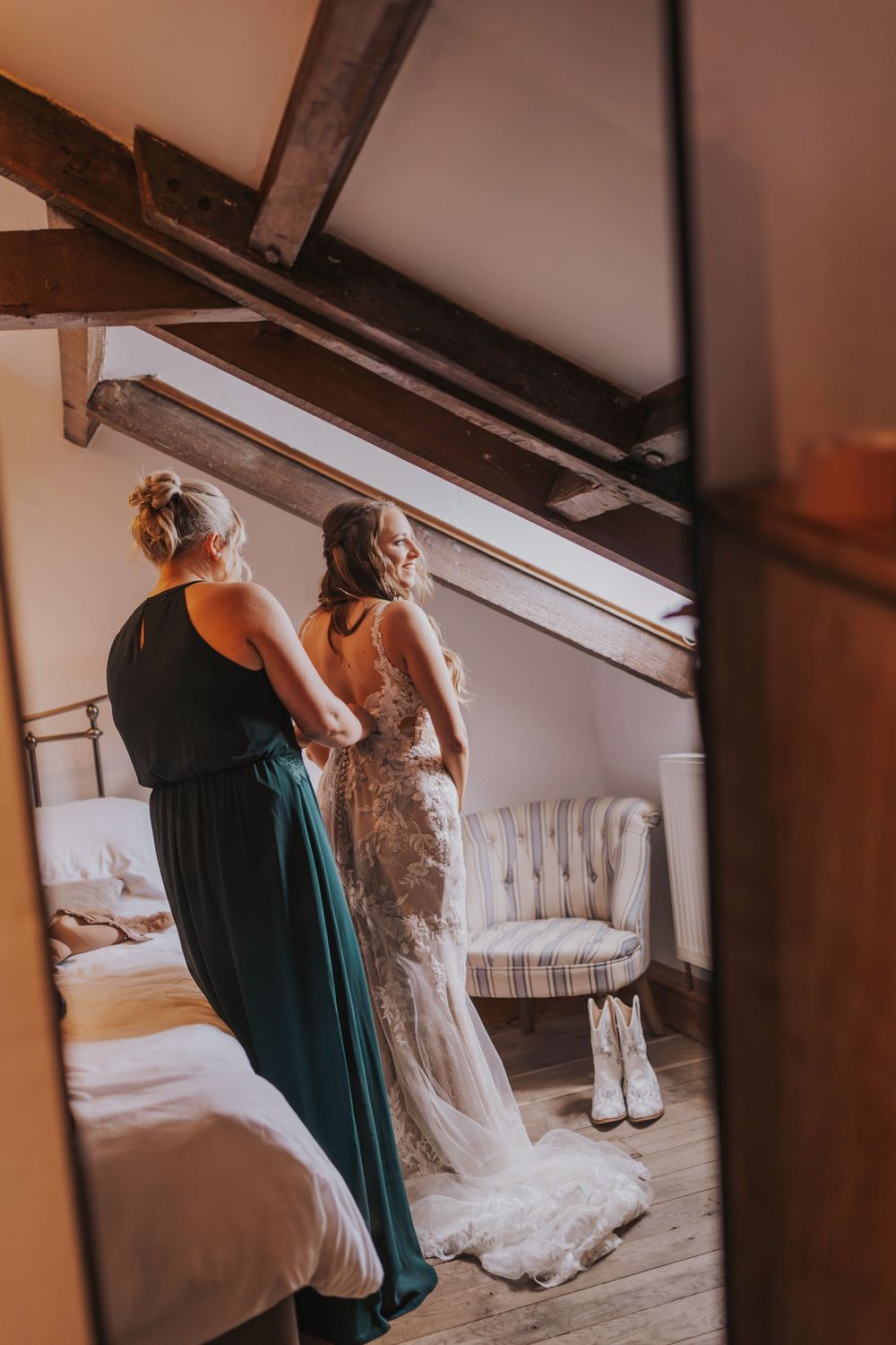 Two women in formal attire help with wedding dress preparations in a rustic attic room.