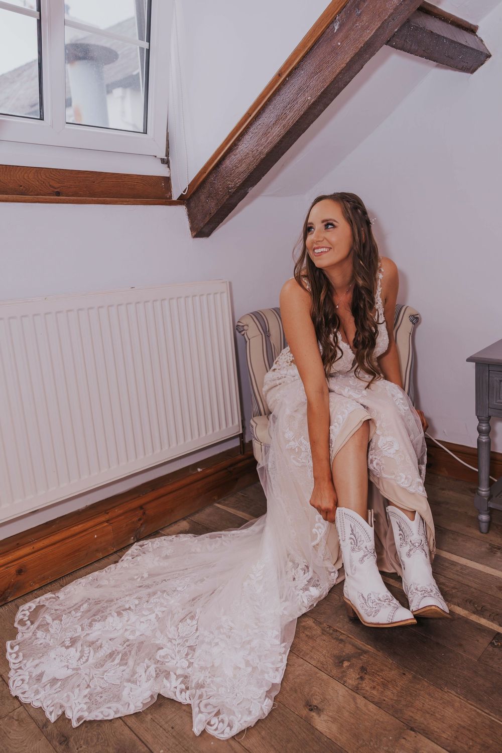 A bride in a white lace wedding dress puts on cowboy boots while sitting in a rustic room with wooden beams.