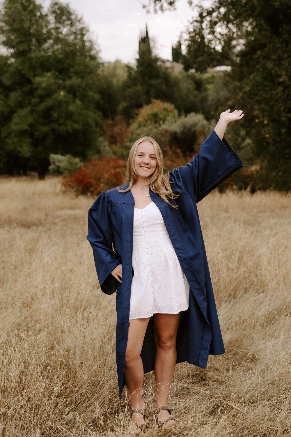 A graduate in a navy blue cap and gown stands joyfully in a golden field on a fall day.