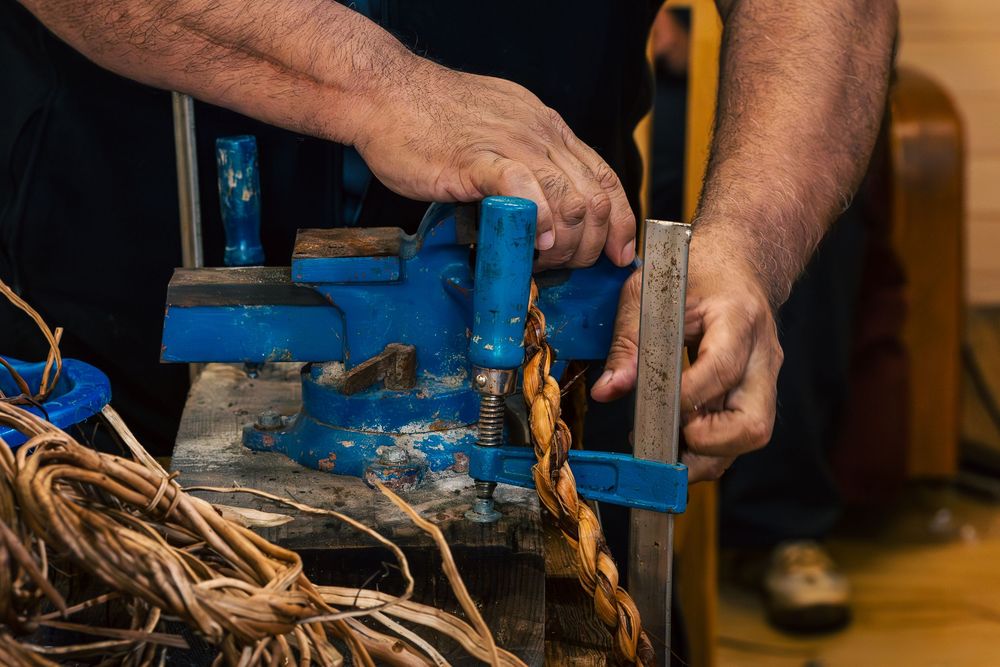 Hands using a blue metal vise to work with natural fiber rope or twine on a workbench.