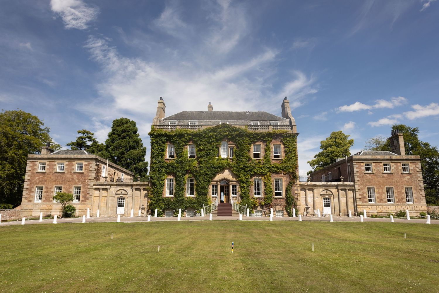 A panoramic view of a historic Georgian manor house with ivy-covered walls and manicured lawns under a blue sky.