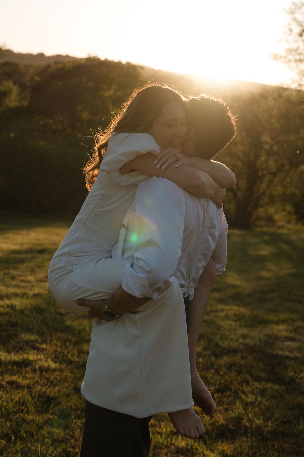 Two people embrace in a grassy field during golden hour sunset with sunlight creating a warm backlit glow.