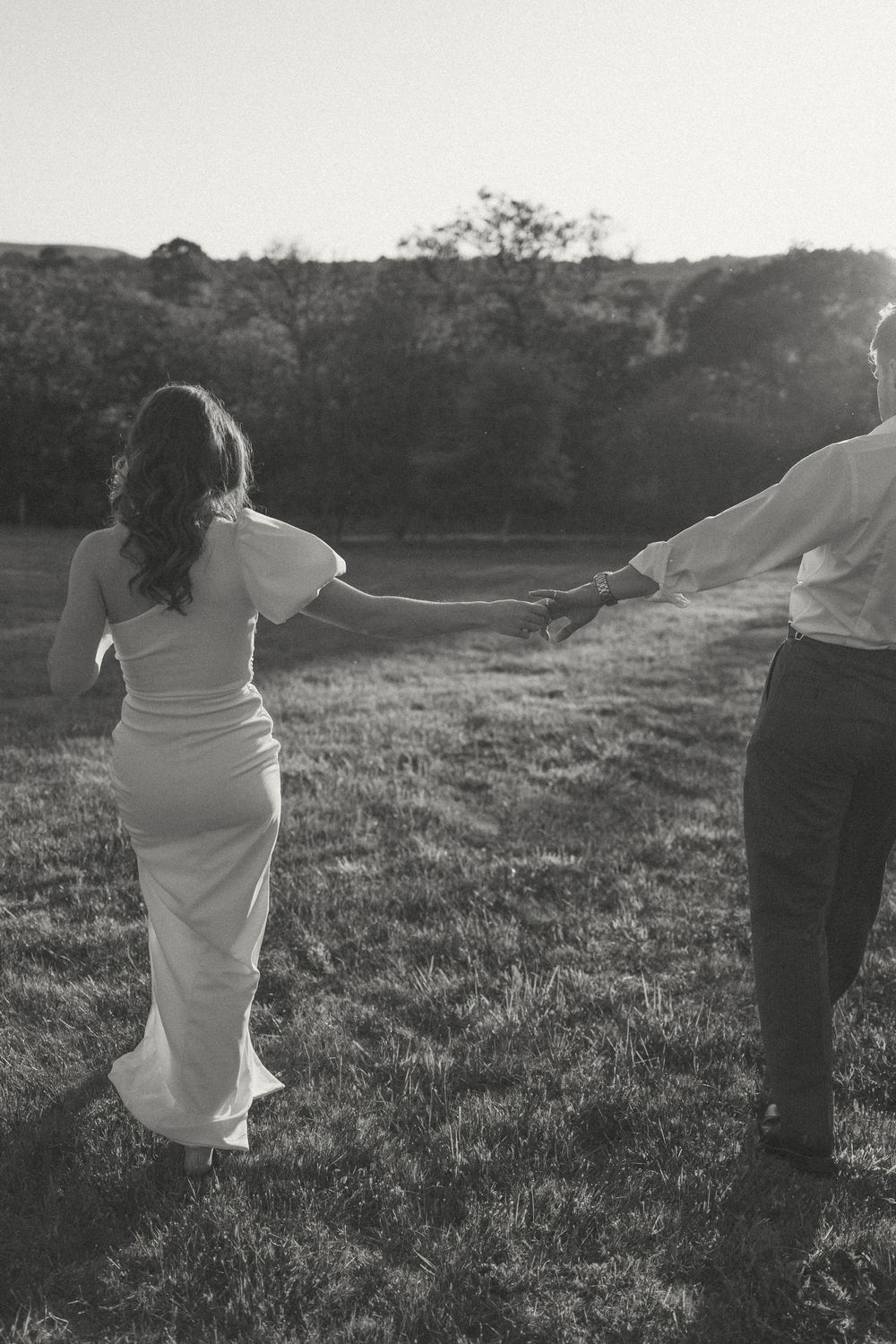 A couple holding hands walks through a grassy field at sunset, with the person in white wearing a long flowing dress.