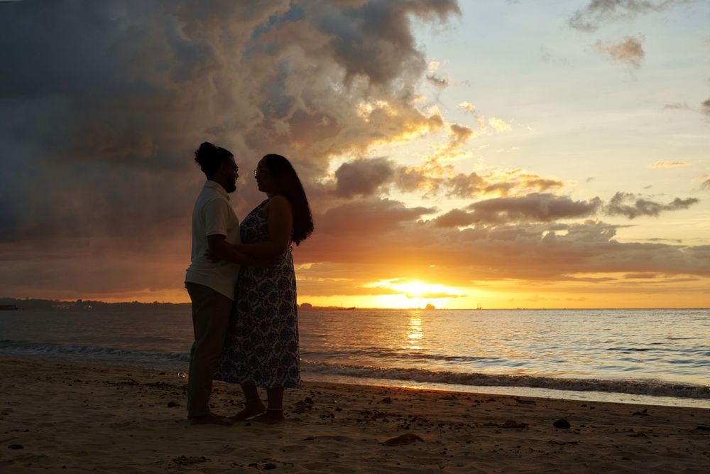 Silhouettes of two people sitting on beach watching dramatic orange and purple sunset over ocean waves.