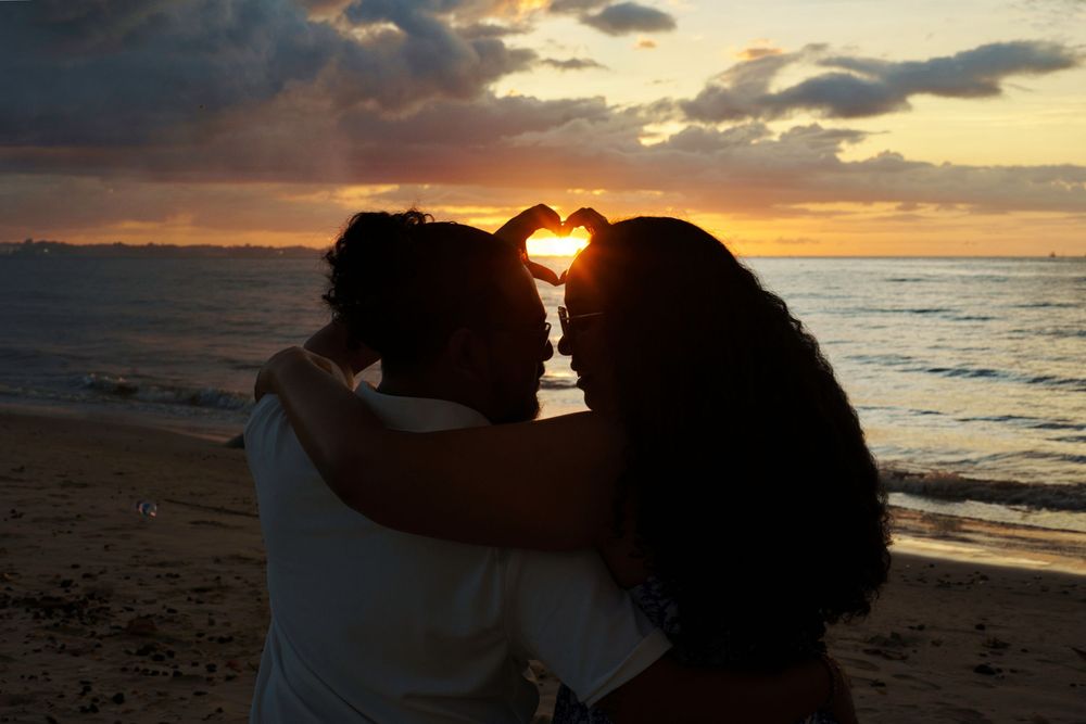 Two figures create heart shape with hands against vibrant golden sunset sky at tropical beach.