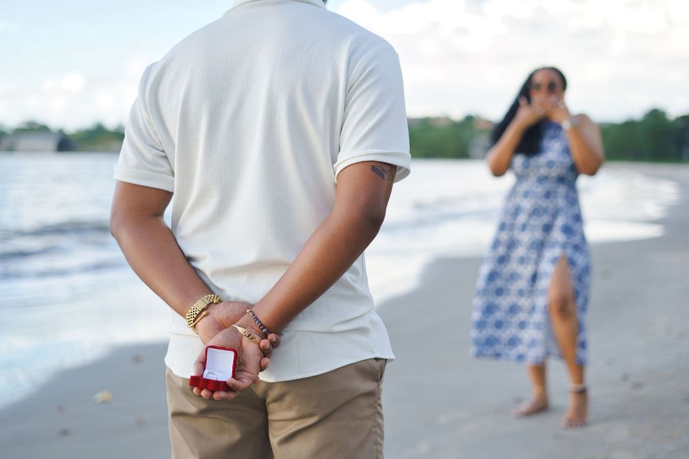 Person holding engagement ring behind back while another in blue dress stands on beach during surprise proposal.