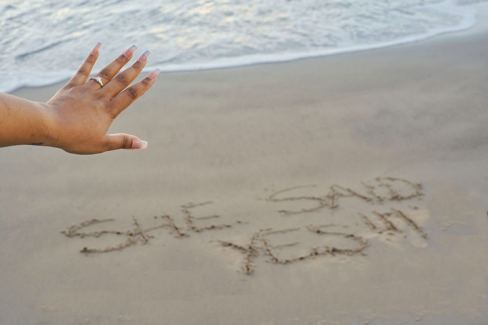 A hand reaches over text written in sand that says "she said yes" on a beach by the ocean.