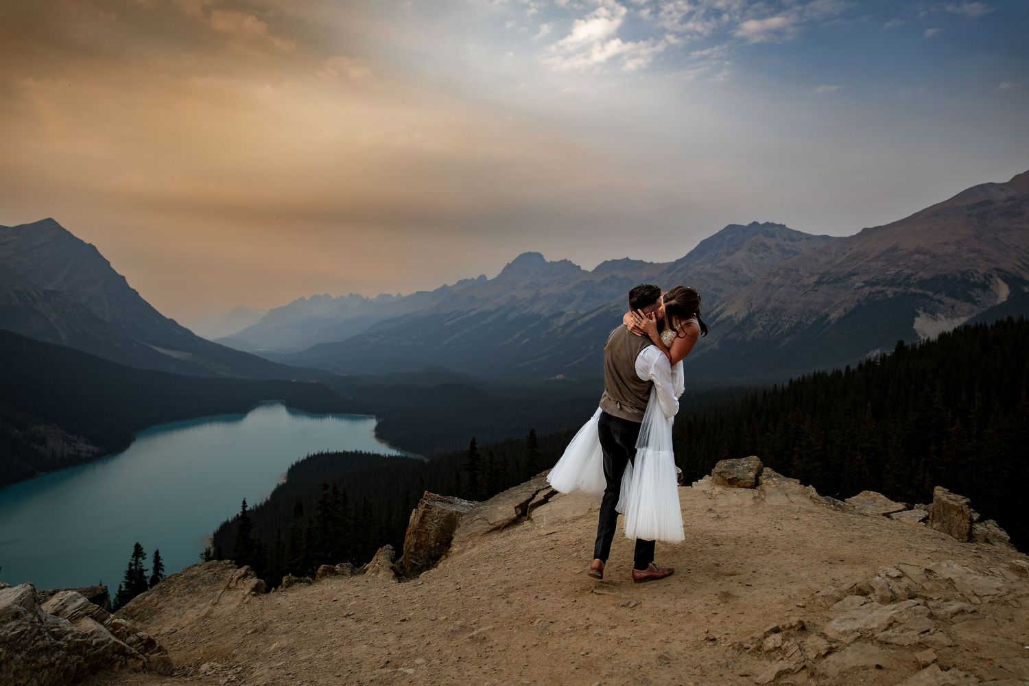 A couple embraces on a mountain cliff overlooking Peyto Lake in Banff National Park during a dramatic sunset.