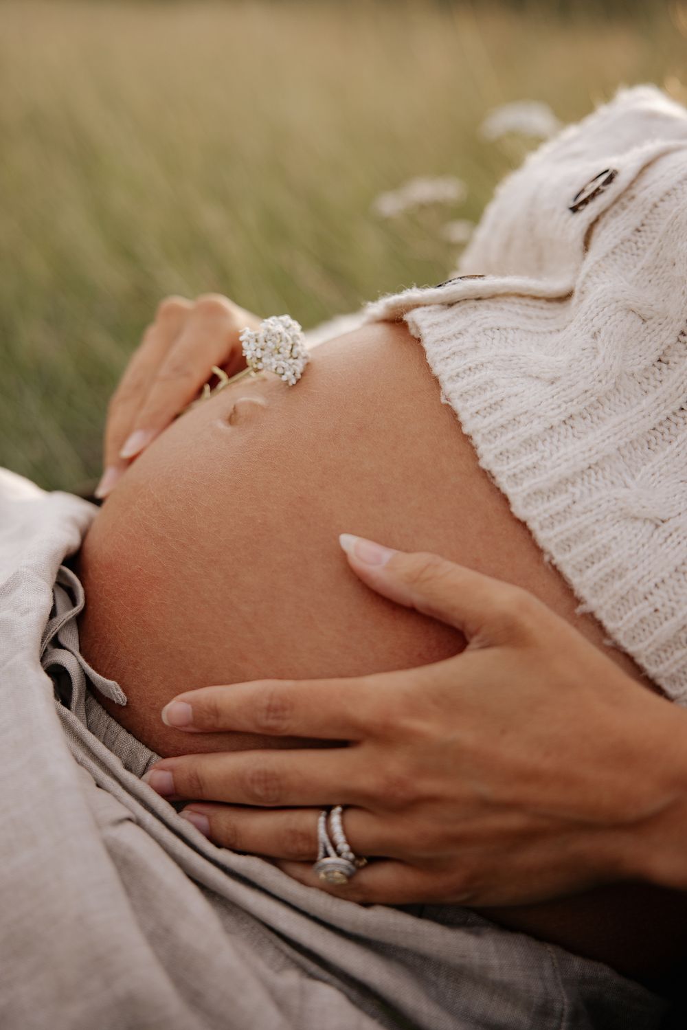 Close-up of hands with engagement ring resting on a pregnant belly wearing a white knit sweater in outdoor setting.