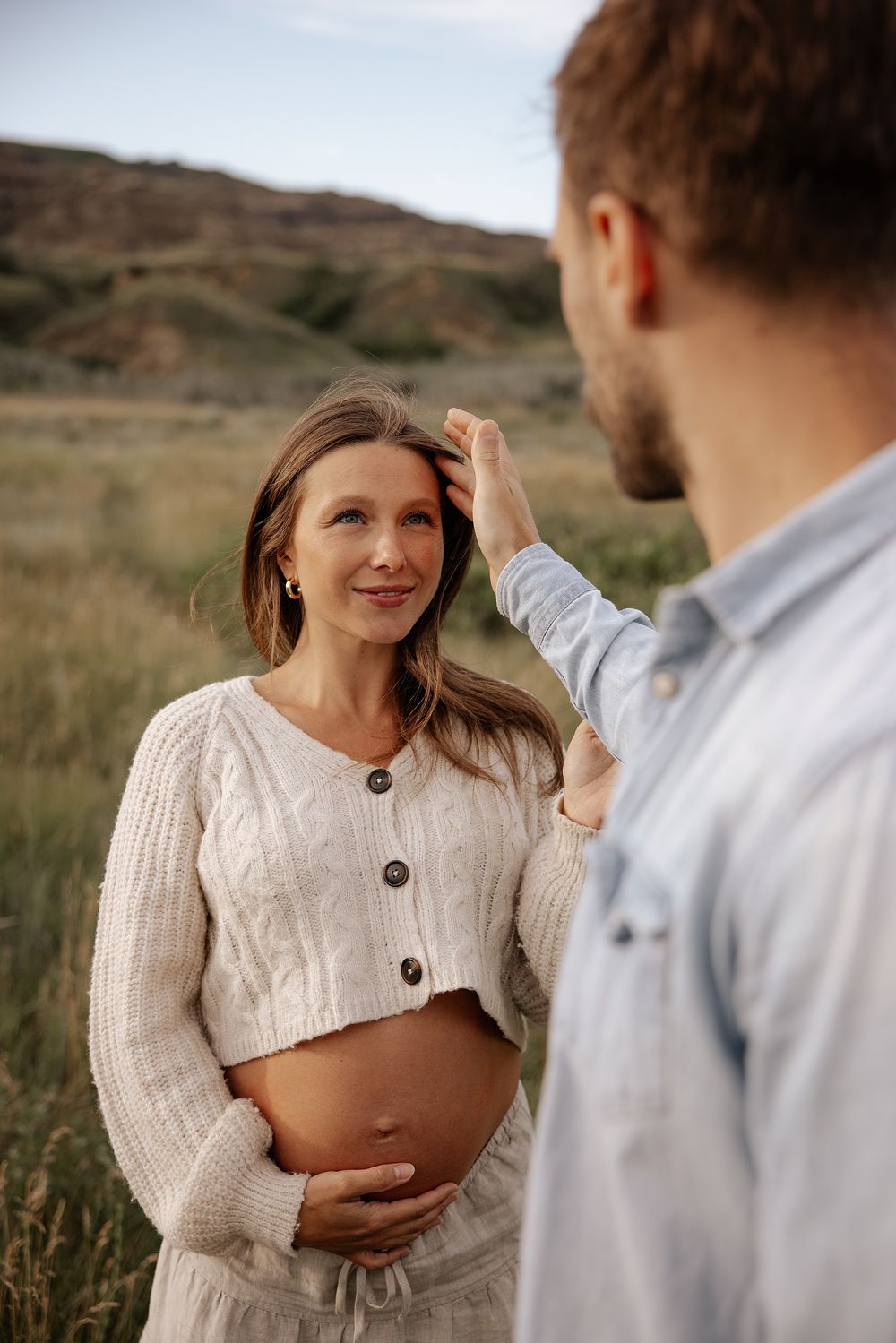 Couple shares intimate moment in field with mountains, wearing white knit sweater and touching hair tenderly.