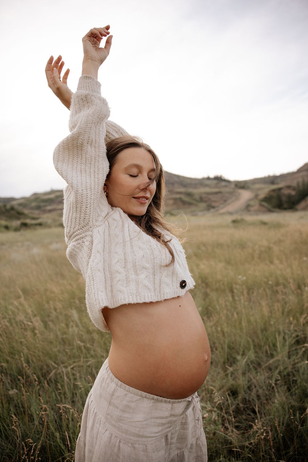 A pregnant woman in a white sweater poses gracefully in a grassy field during a maternity photoshoot.