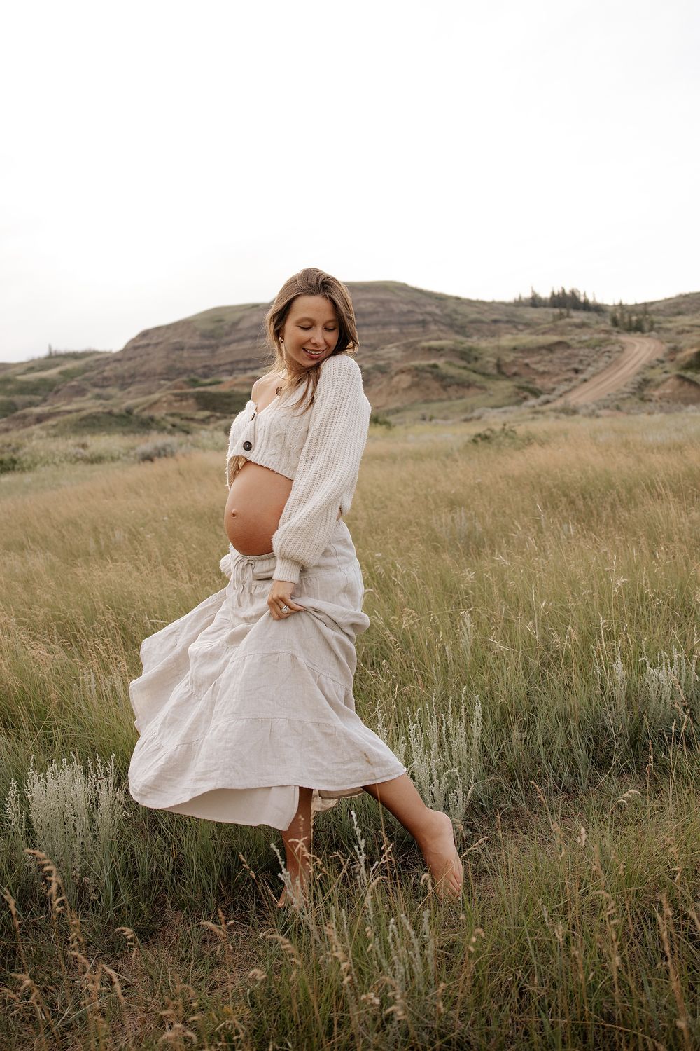 A woman in a flowing white dress dances gracefully through a windswept meadow with rolling hills in the background.