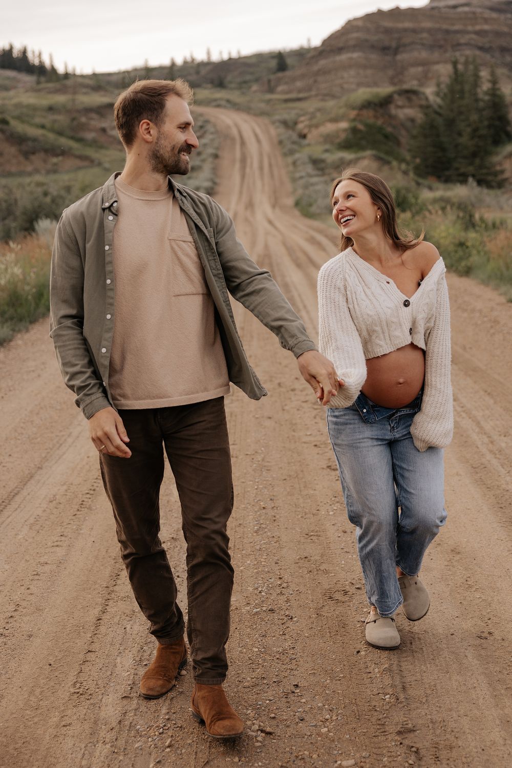 A couple walks hand in hand down a dirt road surrounded by rolling hills and autumn foliage in the countryside.