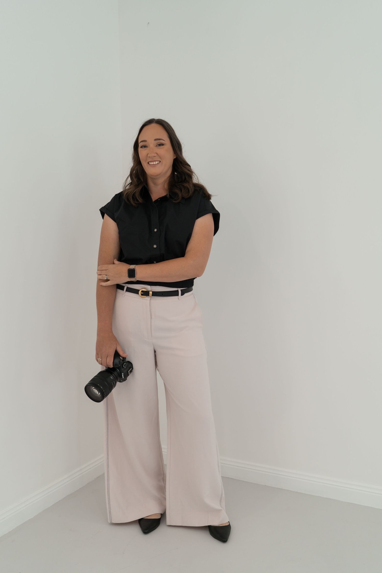Professional photographer in black top and white pants poses in studio against white backdrop with camera in hand.
