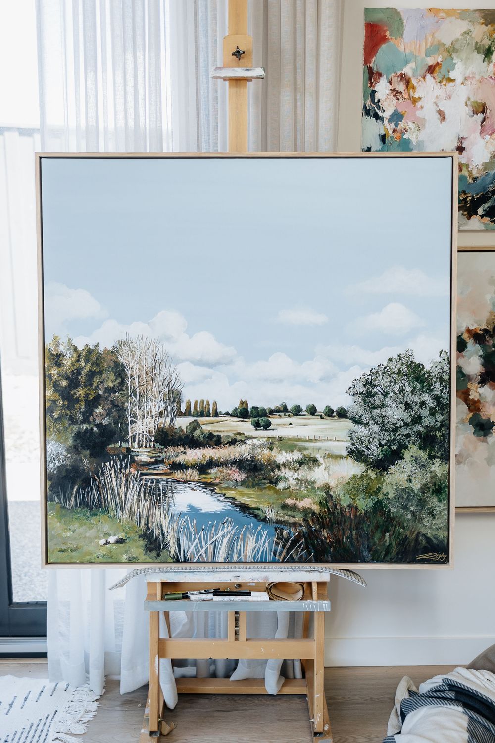 Multiple watercolor landscape paintings displayed on easels in a bright studio space with white curtains.