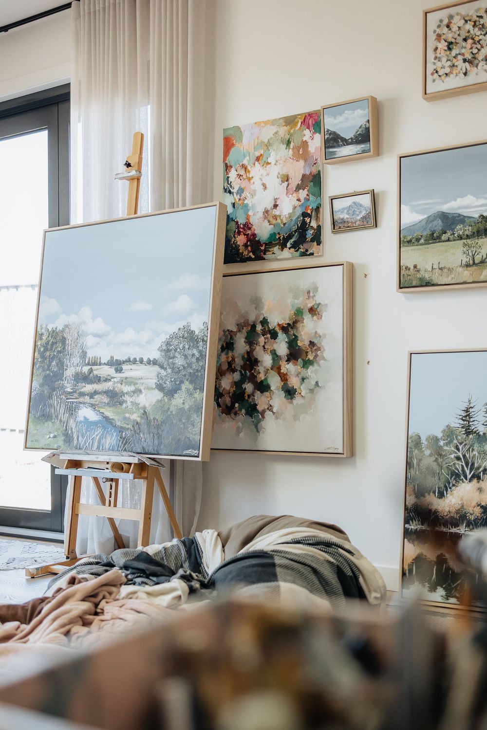 A collection of framed landscape and floral paintings hangs on a bright white gallery wall.