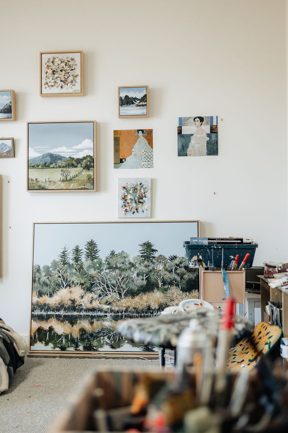 Art studio wall displays landscape paintings and sketches with art supplies and brushes in the foreground.