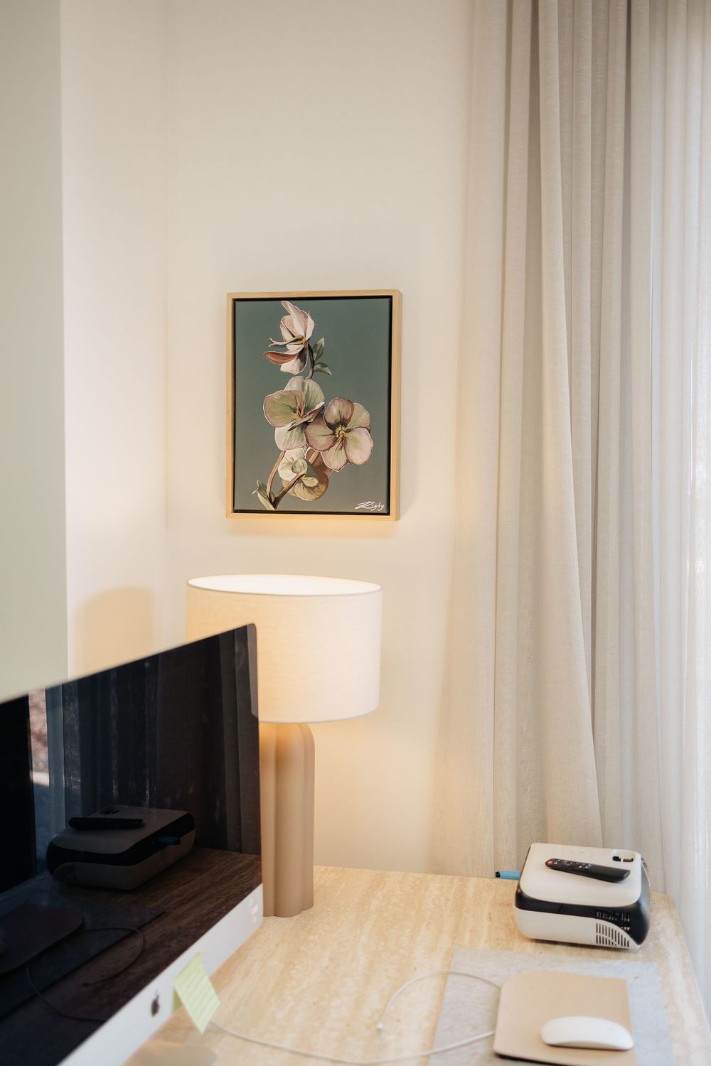 Minimalist bedroom interior with white curtains, table lamp, and framed floral artwork on walls.