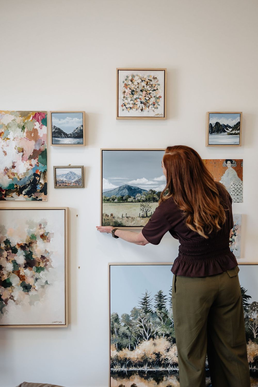 A gallery wall displays various landscape paintings and artwork in simple frames against a white wall.