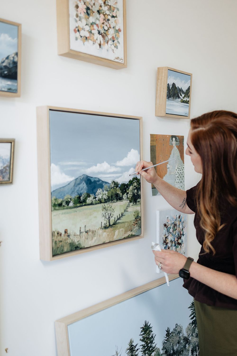 An art gallery wall displays landscape paintings and nature artwork in wooden frames against a white wall.
