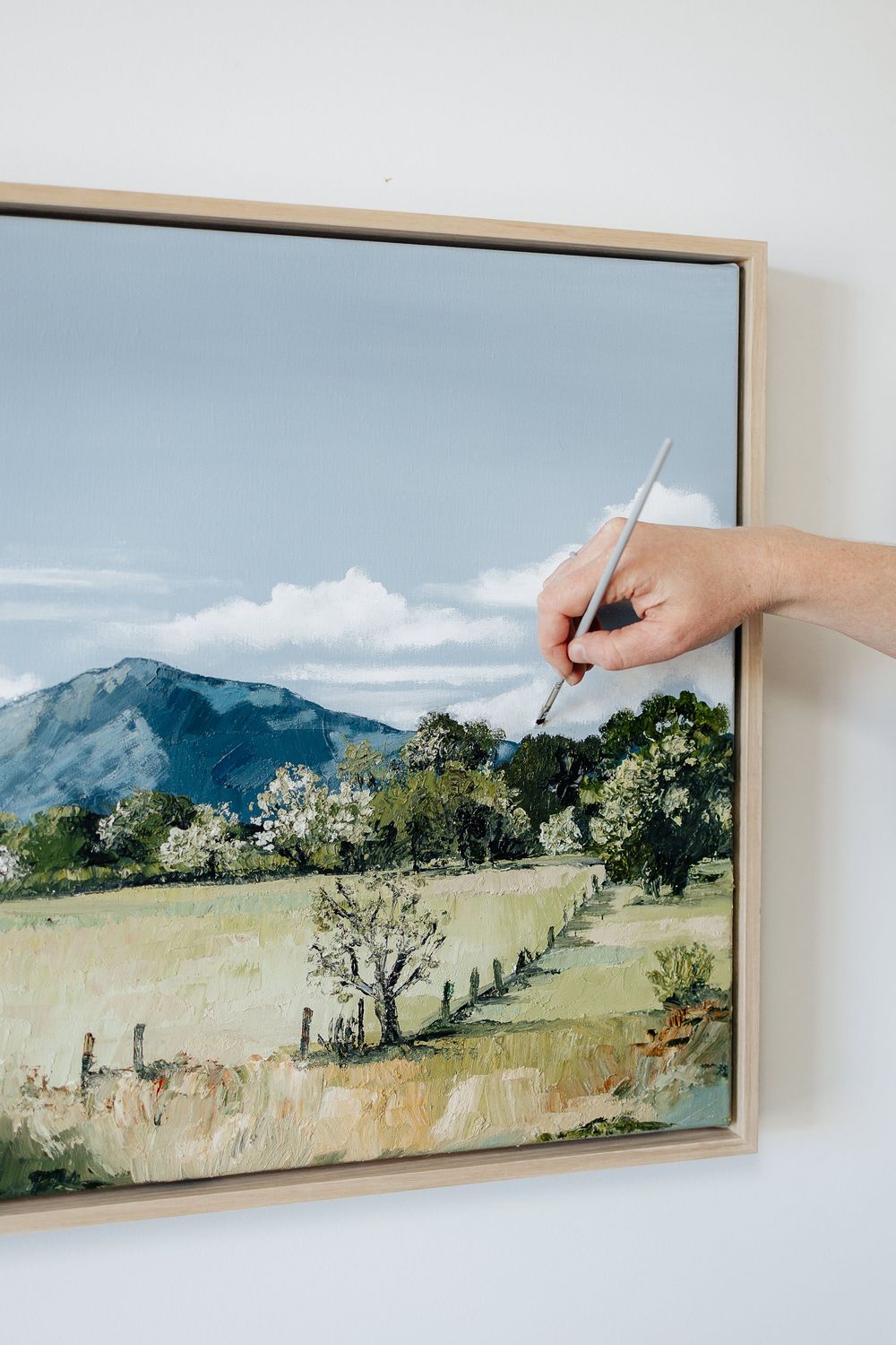 A panoramic landscape painting of rolling hills and trees displayed in a wooden frame with a hand adding details.