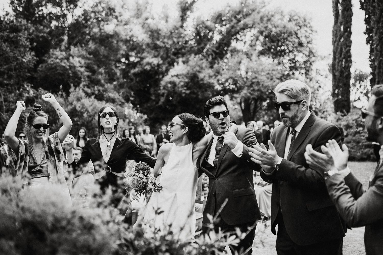 Black and white wedding ceremony scene with guests gathered in garden setting.