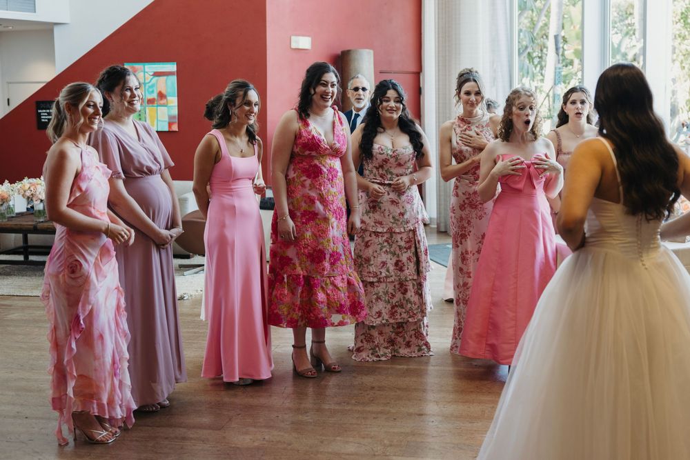 Wedding party wearing various pink and coral dresses gathered in an indoor venue.