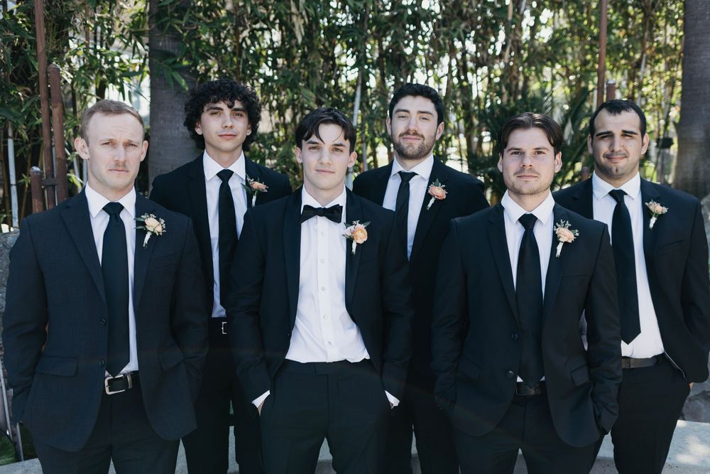 Groomsmen in black suits and ties pose together outdoors among trees.