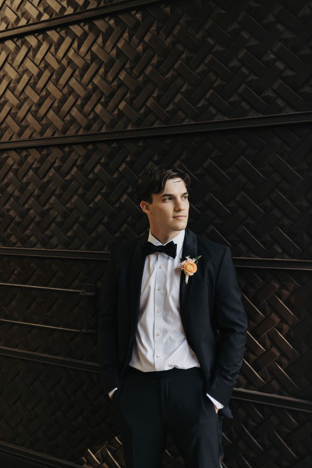 Formal portrait against textured dark wall with boutonniere and classic tuxedo styling.